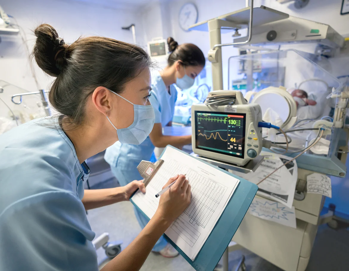 Two nurses in blue scrubs and masks monitoring a newborn in a NICU incubator with medical equipment displaying vital signs